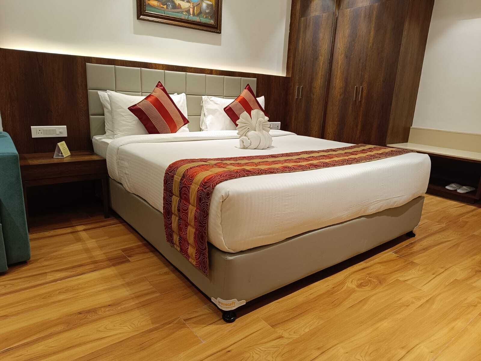 A neatly made bed with a white comforter, red and beige patterned throw, and two white pillows. There is a wooden nightstand with a lamp, a small table with a notepad, and a pair of slippers on the floor.