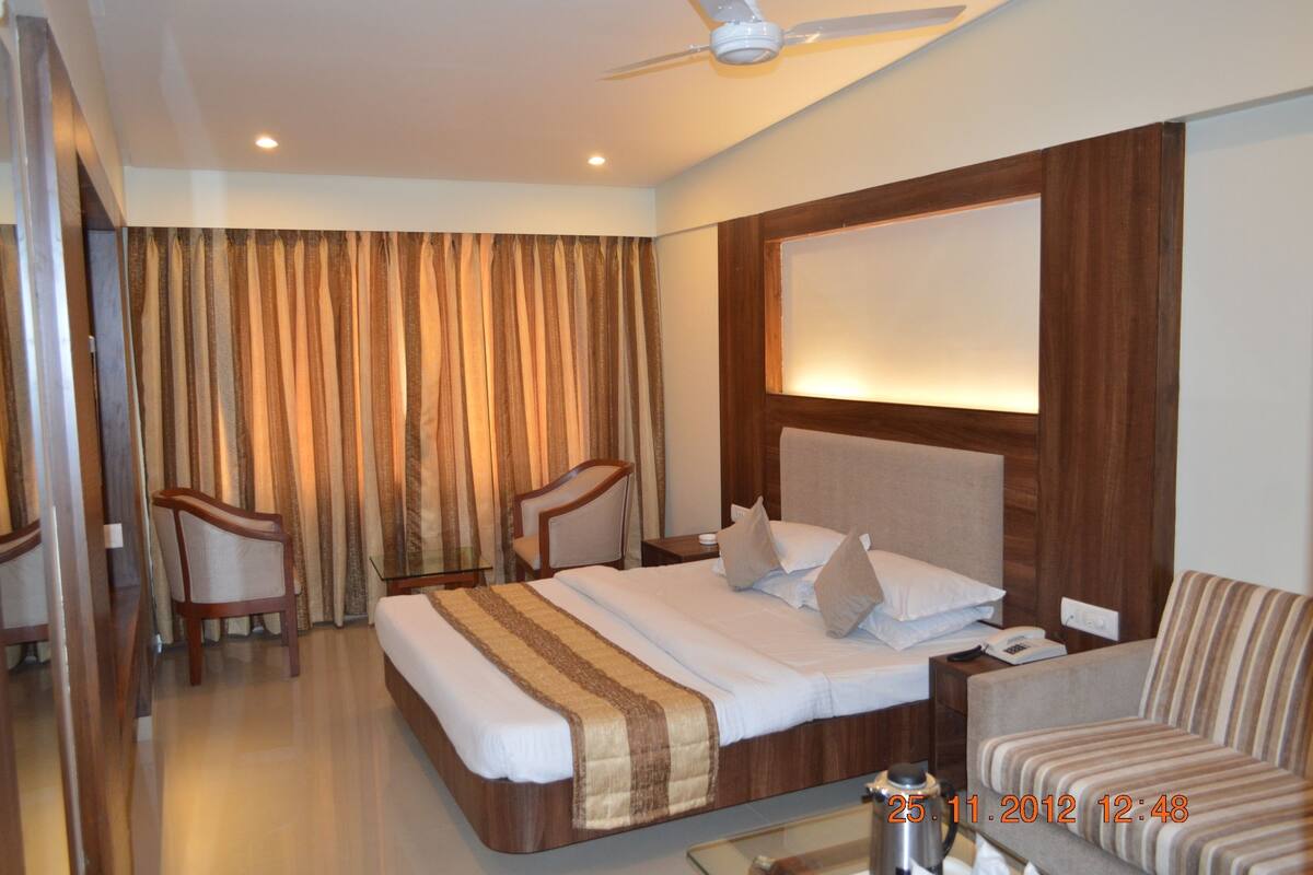 A hotel room with a large bed, a sofa, two armchairs, a ceiling fan, and a large window with curtains.