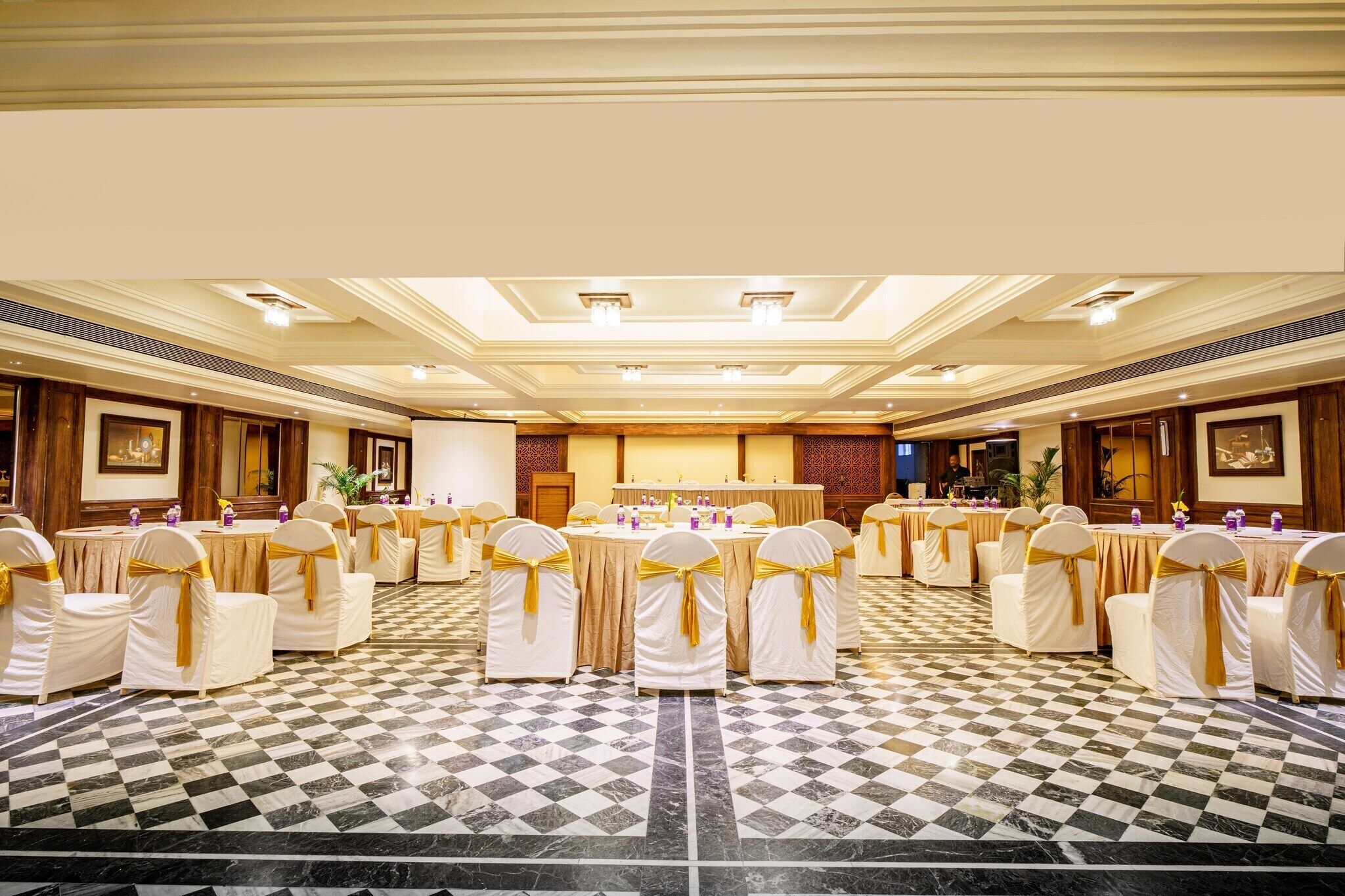 A banquet hall with round tables, chairs with golden sashes, and a checkered marble floor.