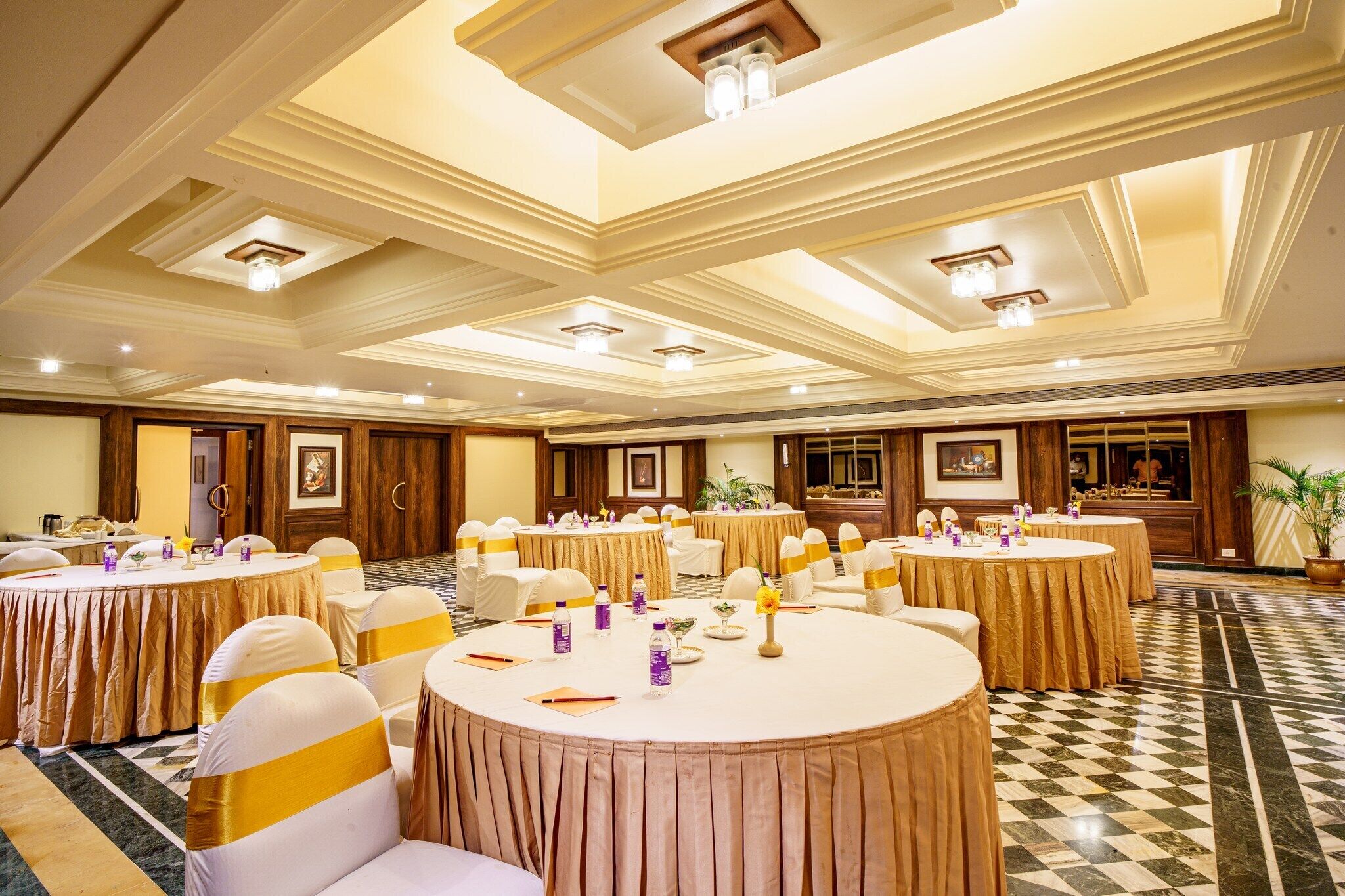 A banquet hall with round tables, white chairs with gold sashes, and a checkered floor.