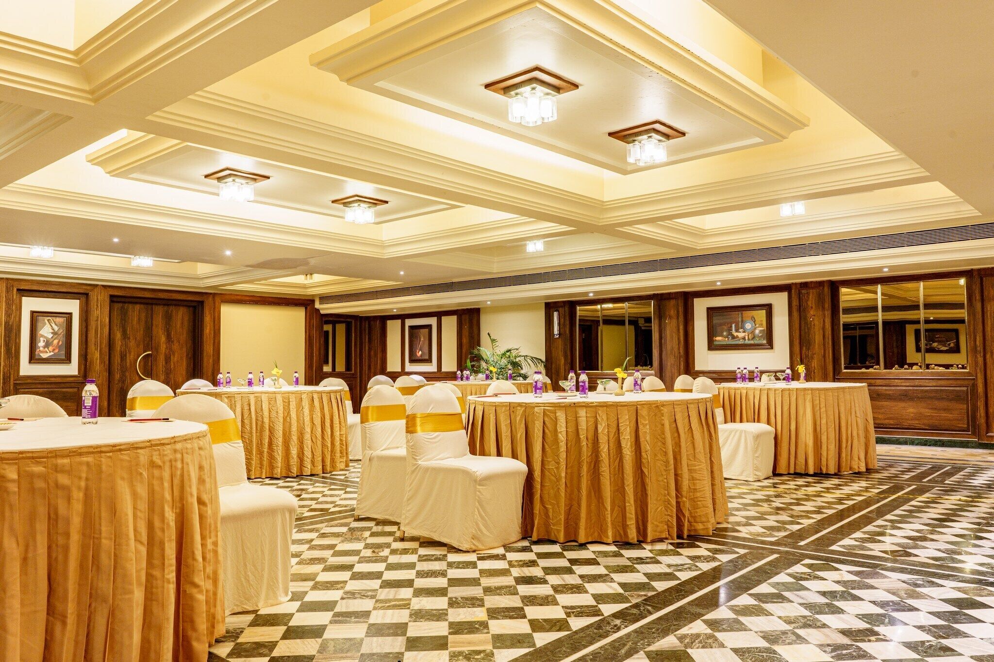 A banquet hall with round tables covered in gold tablecloths and white chairs with gold sashes. The room has a checkered floor and ornate ceiling with chandeliers.