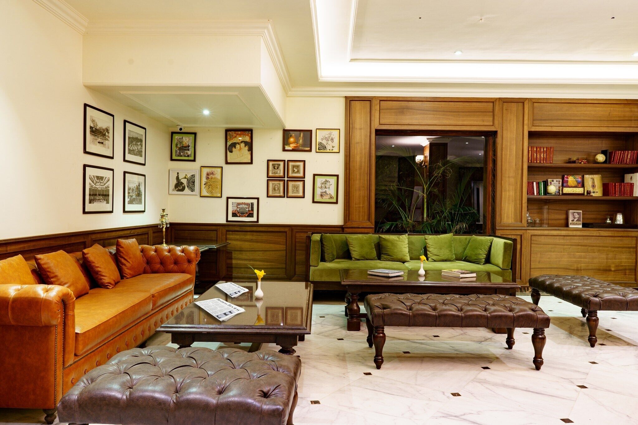 A spacious living room with a leather sofa, a green sofa, and two ottomans. There are framed pictures on the walls and a wooden bookshelf.