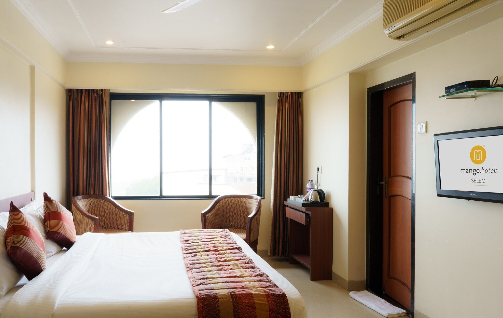 Une chambre d’hôtel avec un lit, deux chaises, un bureau avec un ordinateur, une fenêtre avec des rideaux et un téléviseur fixé au mur.