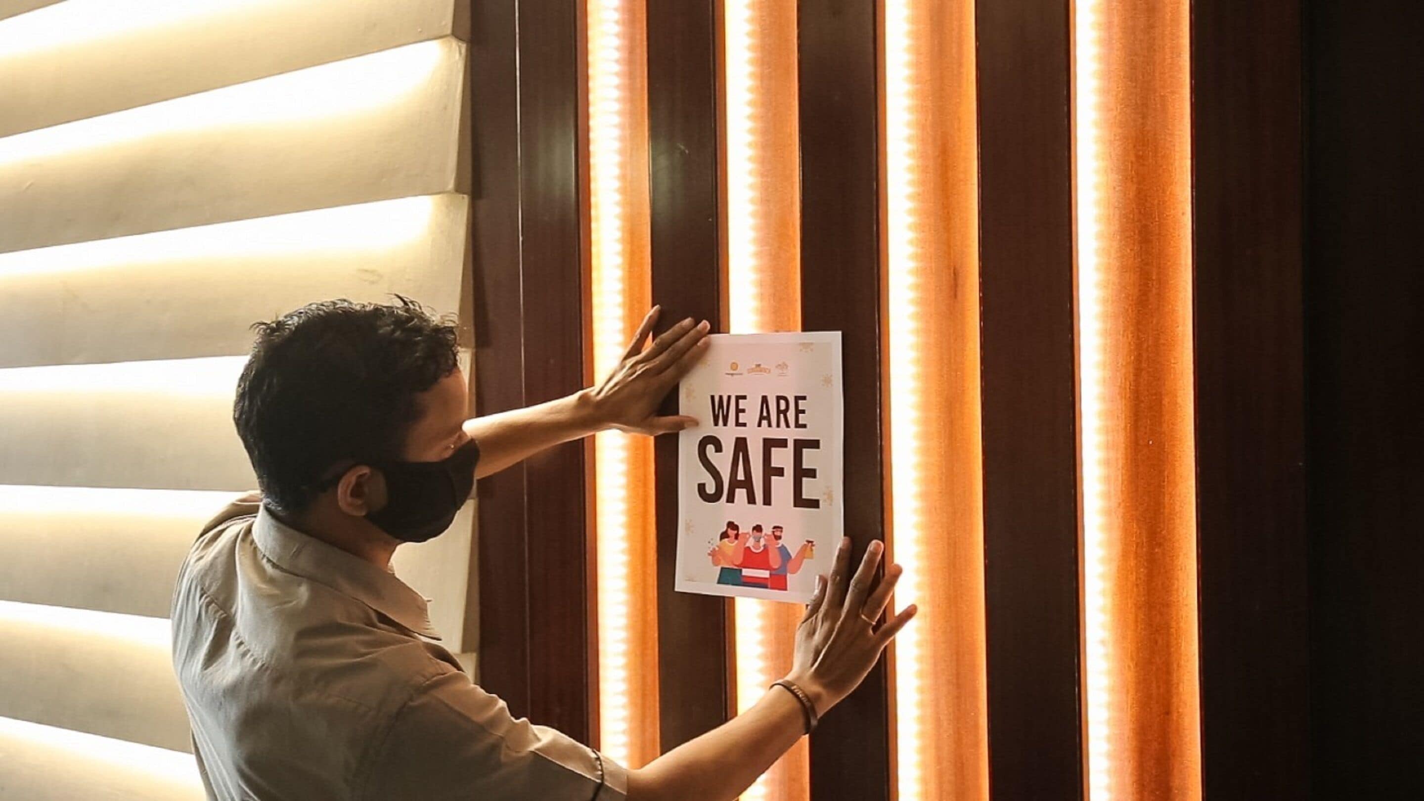 A person holding a sign that reads "WE ARE SAFE" against a wall with vertical wooden slats and a light source behind them.