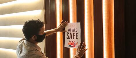 A person holding a sign that reads "WE ARE SAFE" against a wall with vertical wooden slats and a light source behind them.