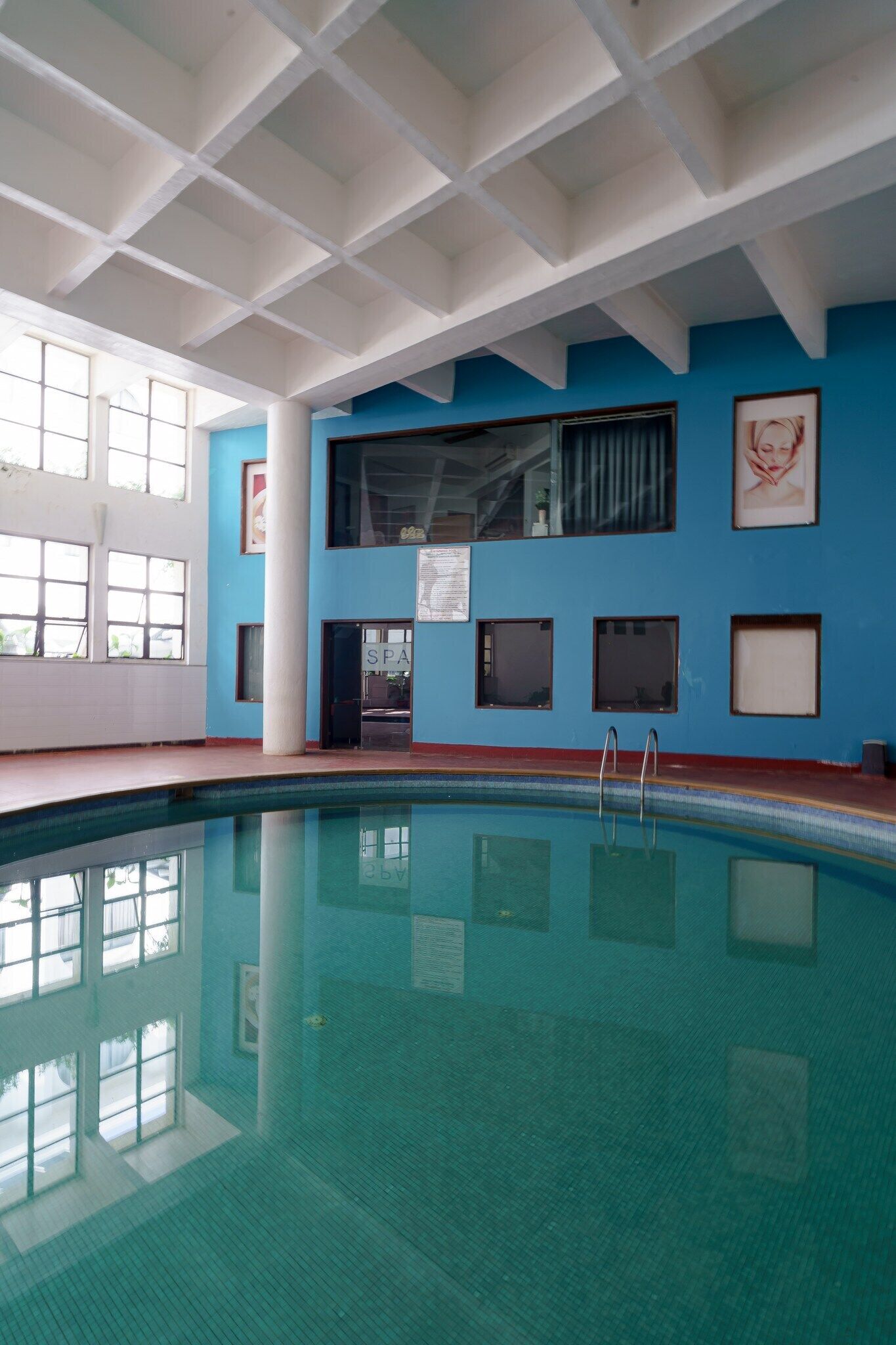 Piscina interior com chão ladrilhado, teto com padrão de grade e parede azul com quadros emoldurados.
