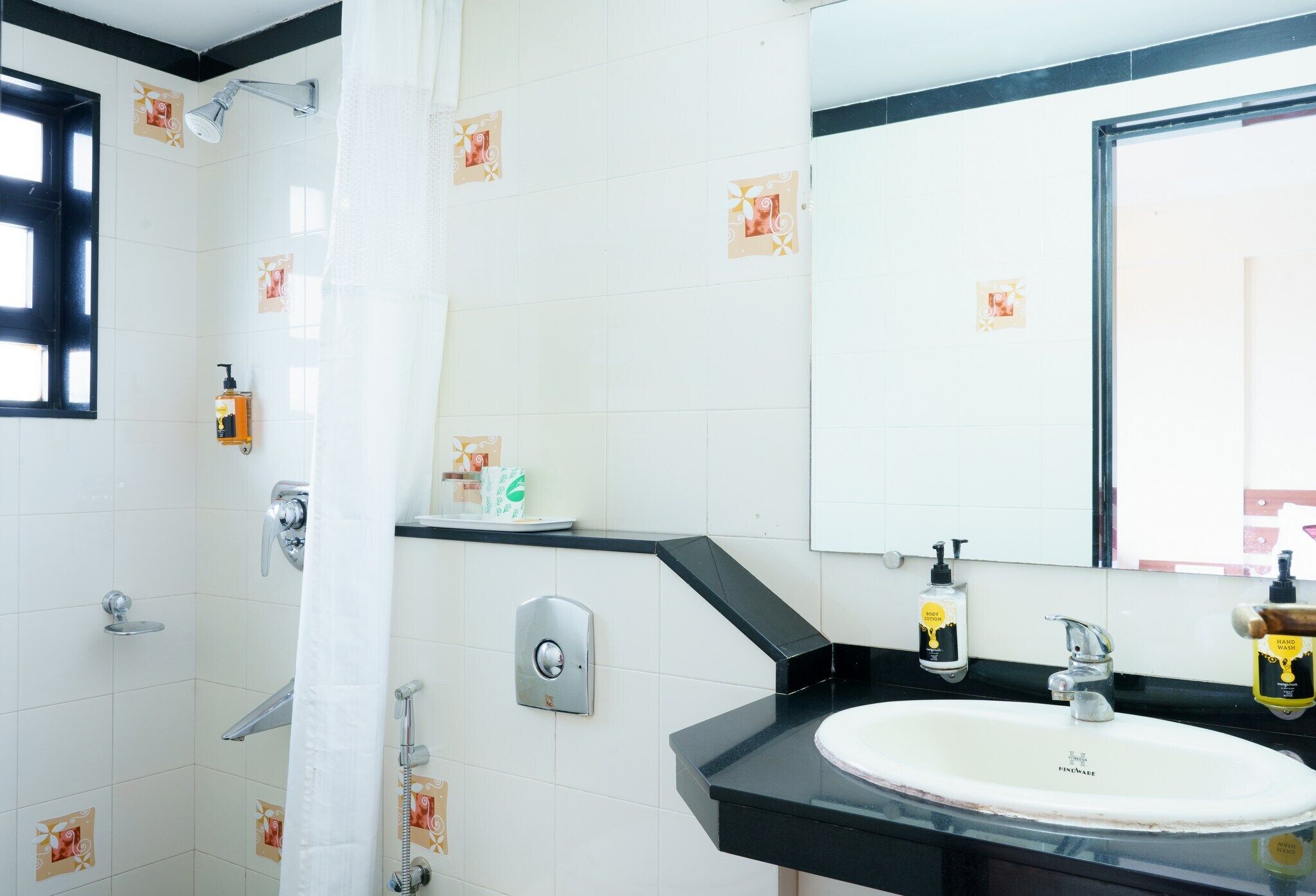 Un baño con un lavamanos blanco, encimera negra y un espejo. Hay una ducha con cortina y un dispensador de jabón en la pared.
