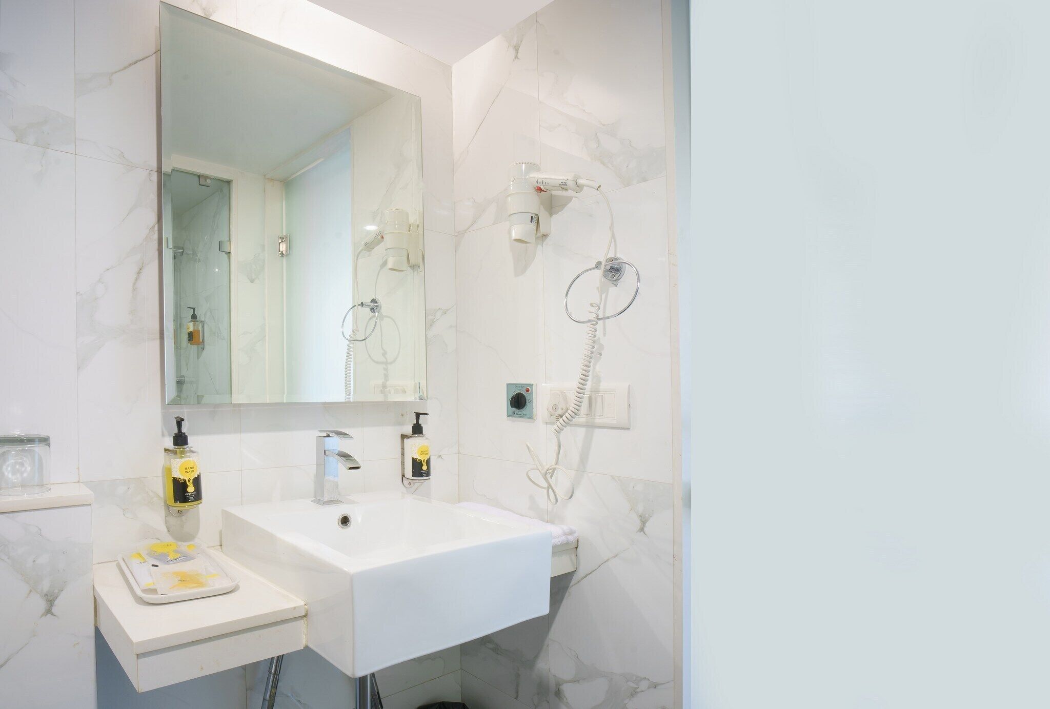 Un baño con lavamanos blanco, un espejo grande, un cabezal de ducha manual y un perchero para toallas.