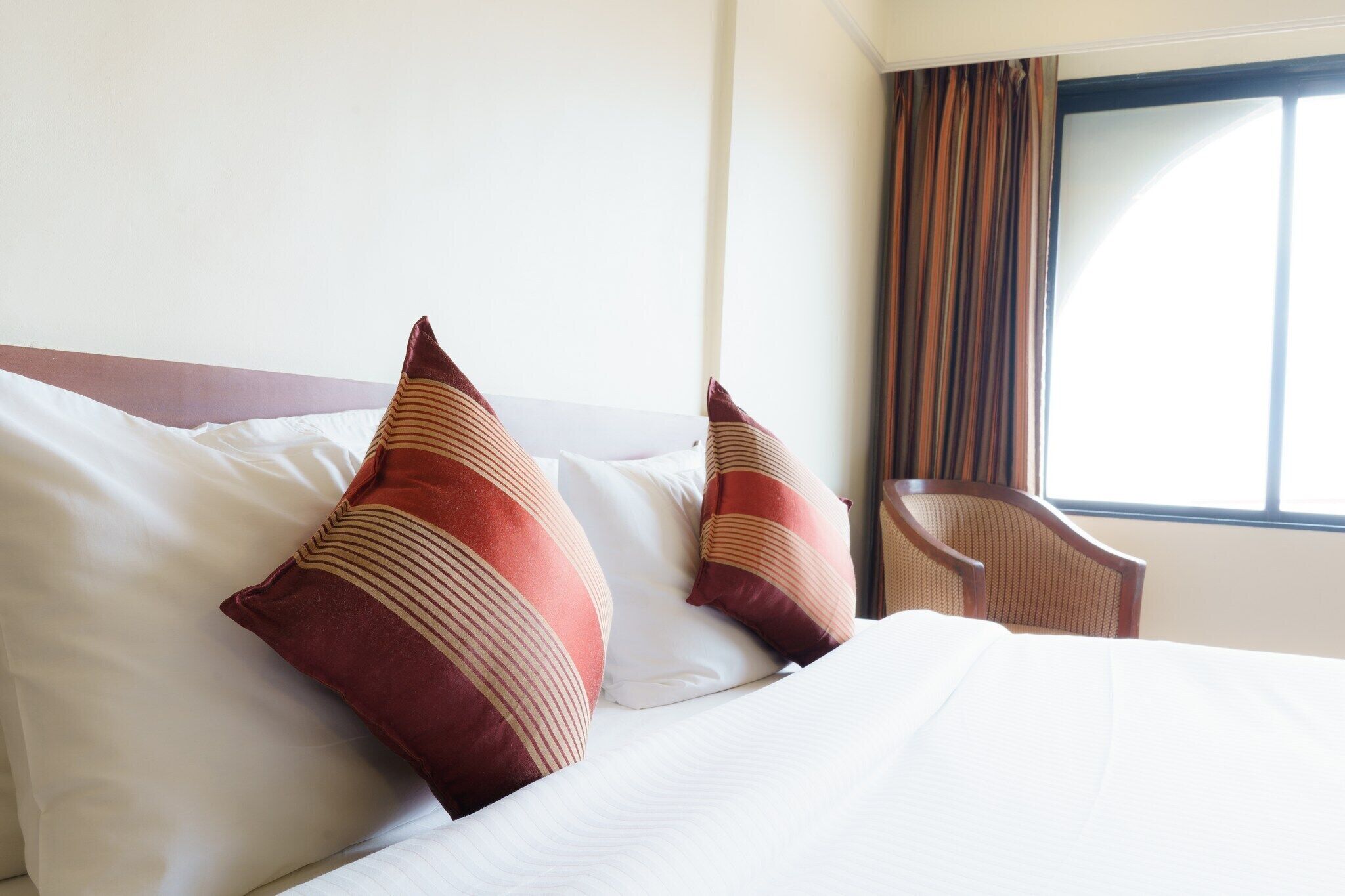 Una habitación de hotel con una cama bien hecha, dos almohadas, una silla y una ventana con cortinas.