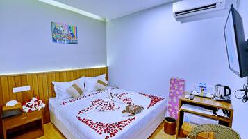 Una cama bien hecha con un arreglo de pétalos de rosa en forma de corazón, una mesita de noche con un abanico y flores, una pequeña mesa con una tetera y tazas, y un televisor montado en la pared.