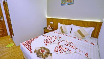 Ein ordentlich bezogenes Bett mit floralem Überwurf und Kissen, ein Nachttisch mit Blumenvase und ein farbenfrohes Bild an der Wand.