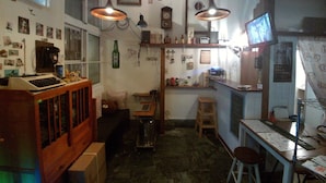 Une petite pièce faiblement éclairée, avec un bureau en bois, une chaise, un petit tabouret et une étagère contenant divers objets. On y trouve deux lampes suspendues, une horloge et un calendrier accrochés au mur.