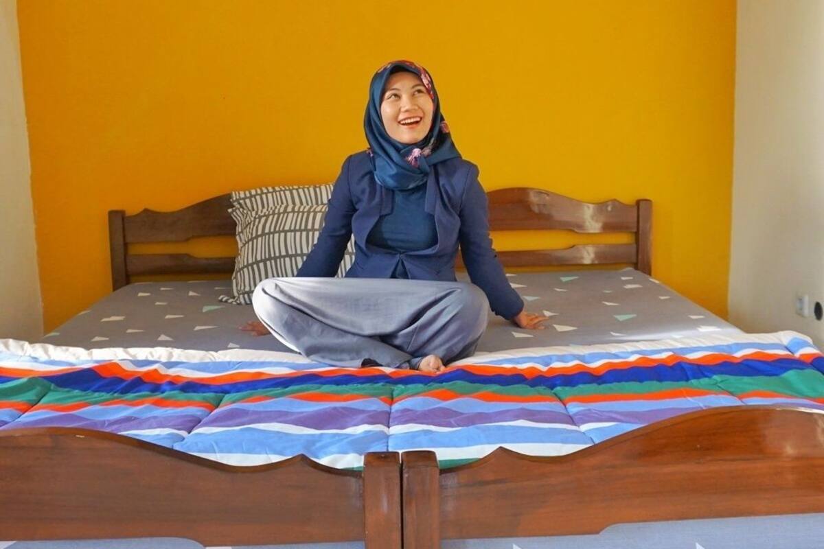 A person sitting on a bed with a colorful striped blanket, a wooden headboard, and a pillow with a striped pattern.