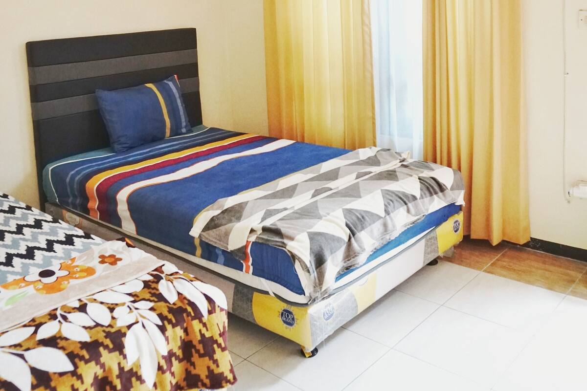 A hotel room with two beds, a large window with yellow curtains, an air conditioning unit, and a tiled floor.