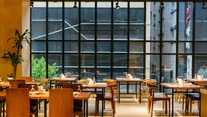 Un restaurante con mesas y sillas de madera, ventanas amplias y vistas a edificios.