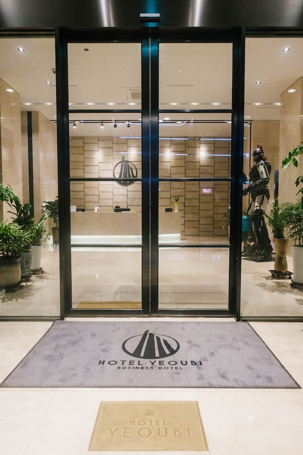 Entrada al Hotel Yeoubi Business Hotel con diseño moderno, que incluye una recepción con pared de azulejos y plantas en macetas.