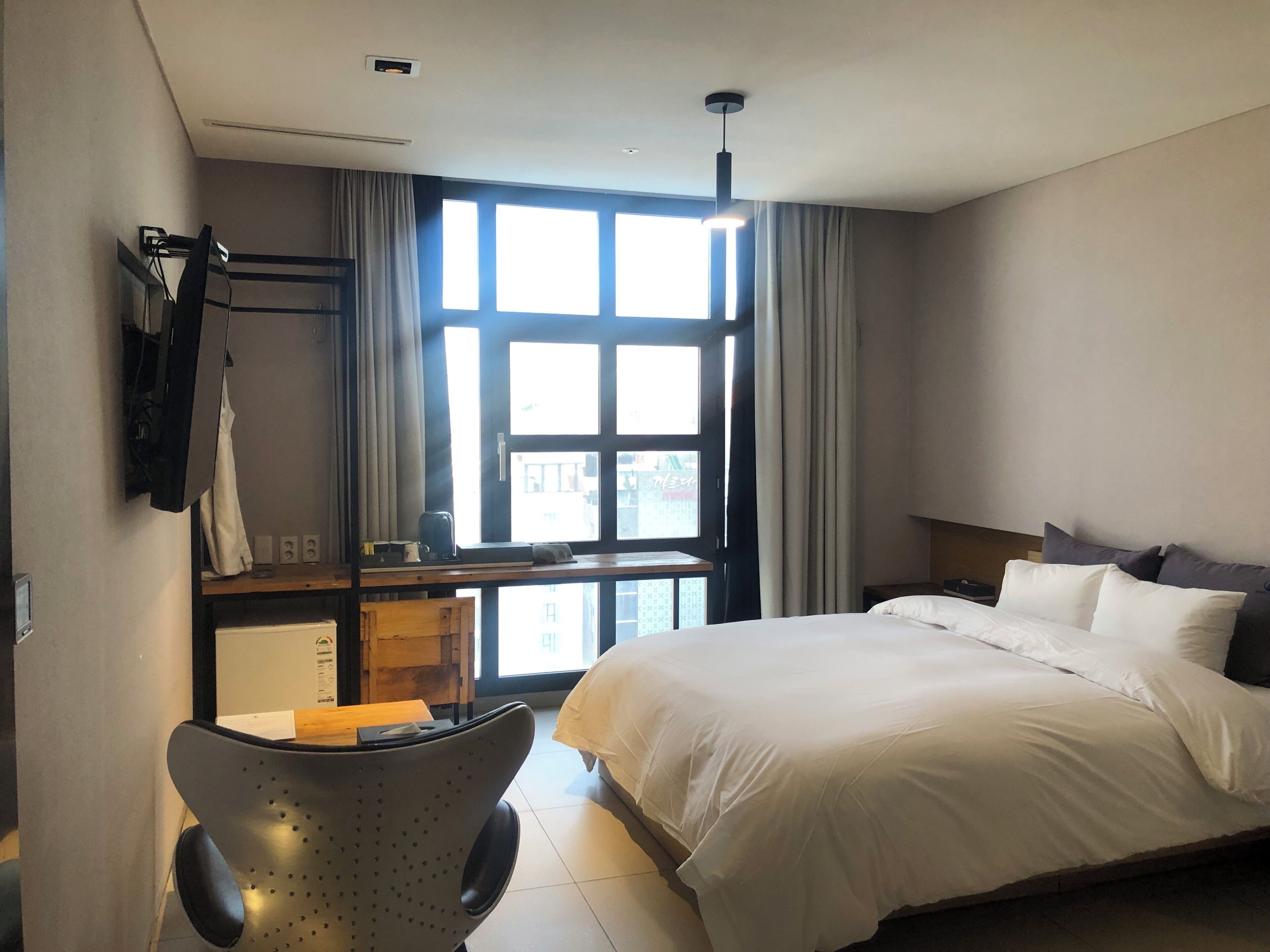 A hotel room with a bed, a desk with a chair, a window with curtains, and a ceiling light.