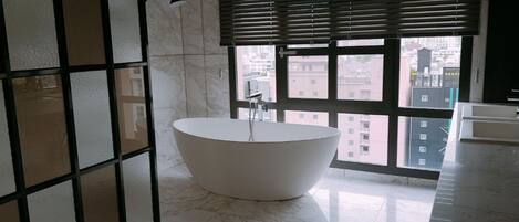 A modern bathroom with a freestanding bathtub, large windows with blinds, and a city view.