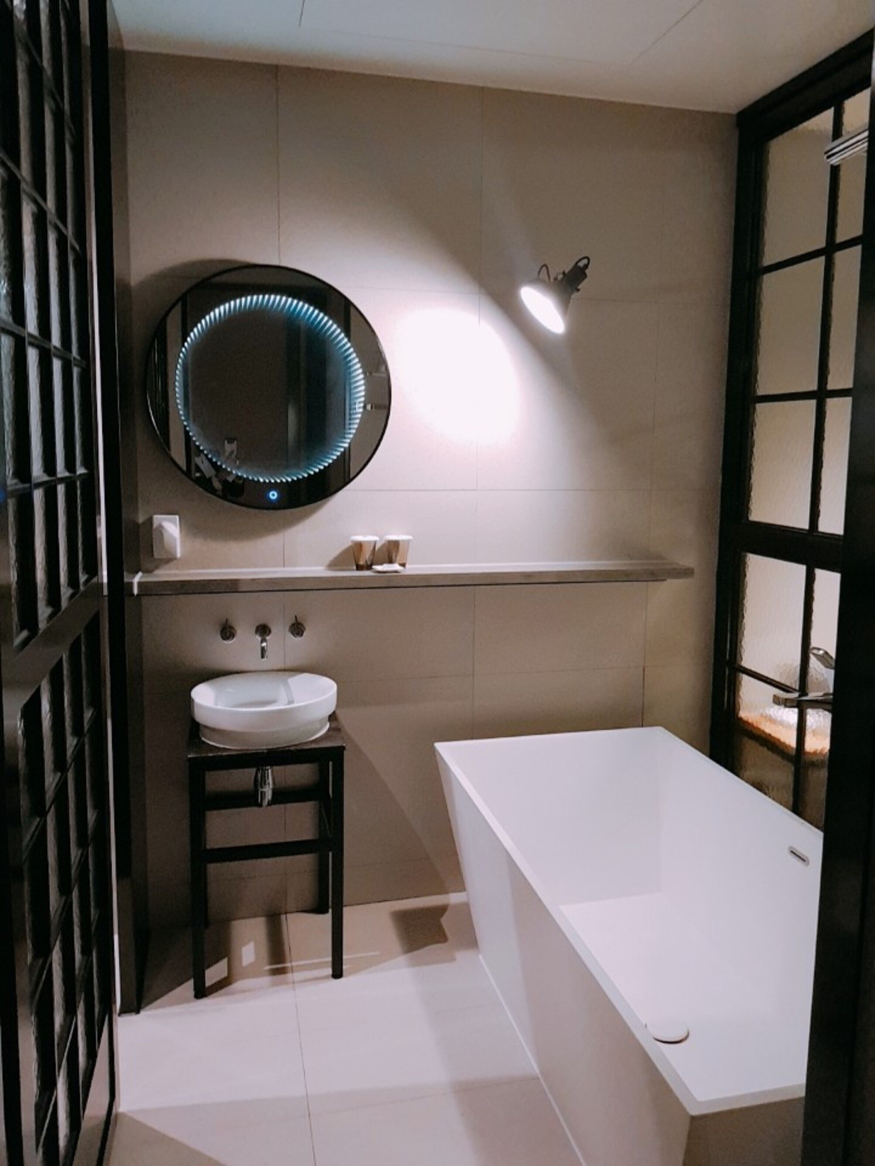 Un baño moderno con un lavamanos blanco, un espejo redondo con luces LED, una bañera blanca y un inodoro.