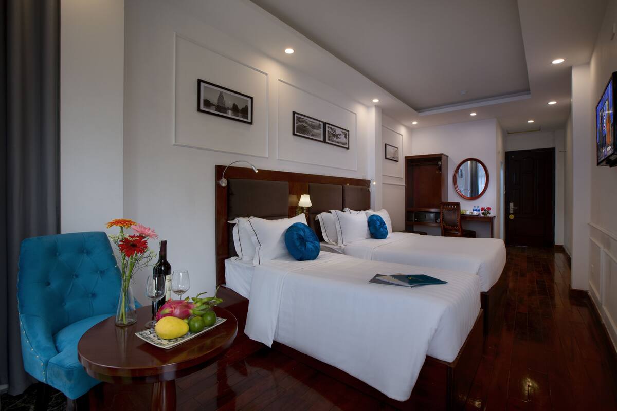 A hotel room with two beds, a blue armchair, a small table with a fruit bowl and a vase, and a mirror on the wall.