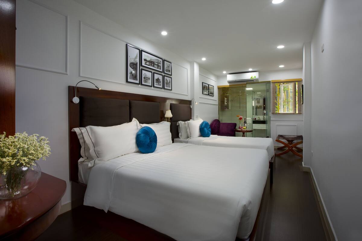 A hotel room with two beds, a bedside table with a vase of flowers, a mirror, and a window with blinds.