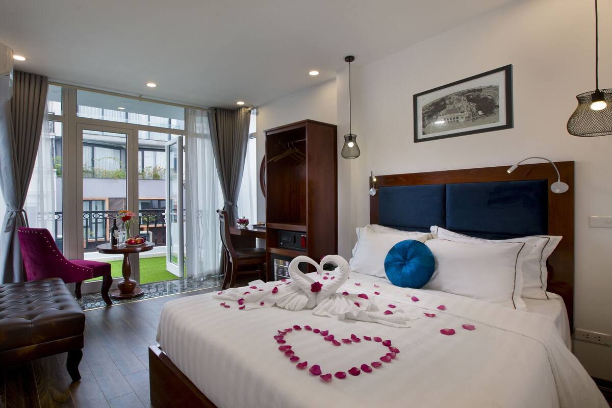 A hotel room with a large bed, a wooden wardrobe, a desk, and a balcony with a chair.