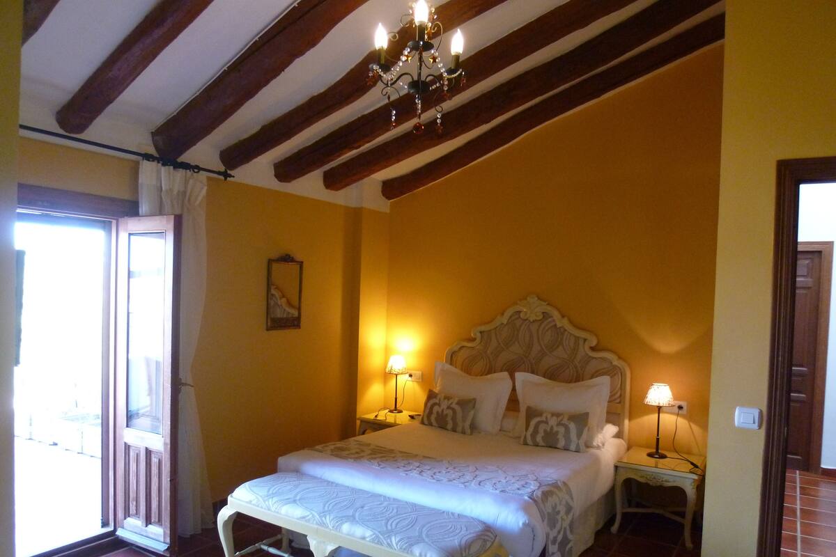 Een slaapkamer met een bed, nachtlampjes, een kroonluchter en een deur naar een balkon.
