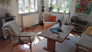 Een gezellige woonkamer met een houten tafel, rieten stoelen en een bank. Twee ramen met gordijnen, een radiator en een boekenkast met boeken en een radio.