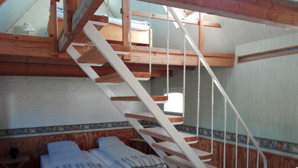 Een kamer met een houten trap die naar een hoger niveau leidt, twee eenpersoonsbedden met witte lakens, een kleine tafel en een raam.