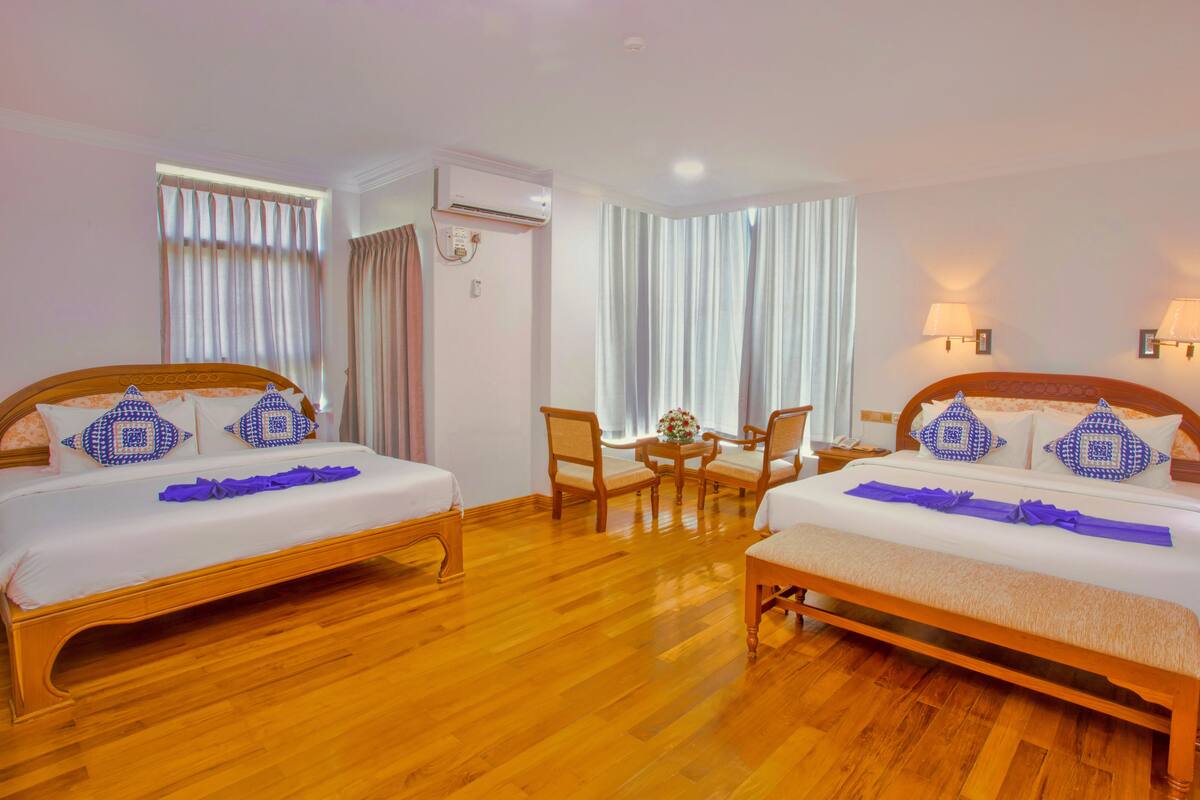 A hotel room with two beds, a wooden floor, a small table with chairs, and a window with curtains.