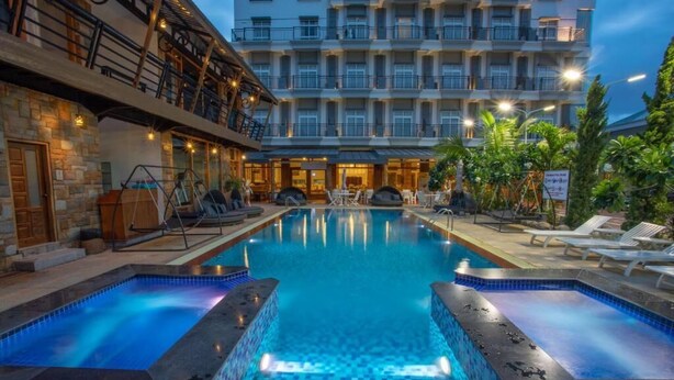 Zona de piscina hotelera con vista despejada, sillas de descanso y un edificio con varios balcones.