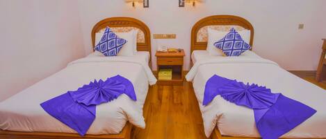 Una habitación con dos camas de madera, cada una con cabecero, sábanas blancas y almohadas decorativas moradas. Hay lámparas fijadas a la pared sobre cada cama y una mesita de noche entre ellas.