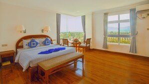 Un dormitorio amplio con una cama grande, un banco de madera, una pequeña mesa con sillas y vistas al verdor por la ventana.