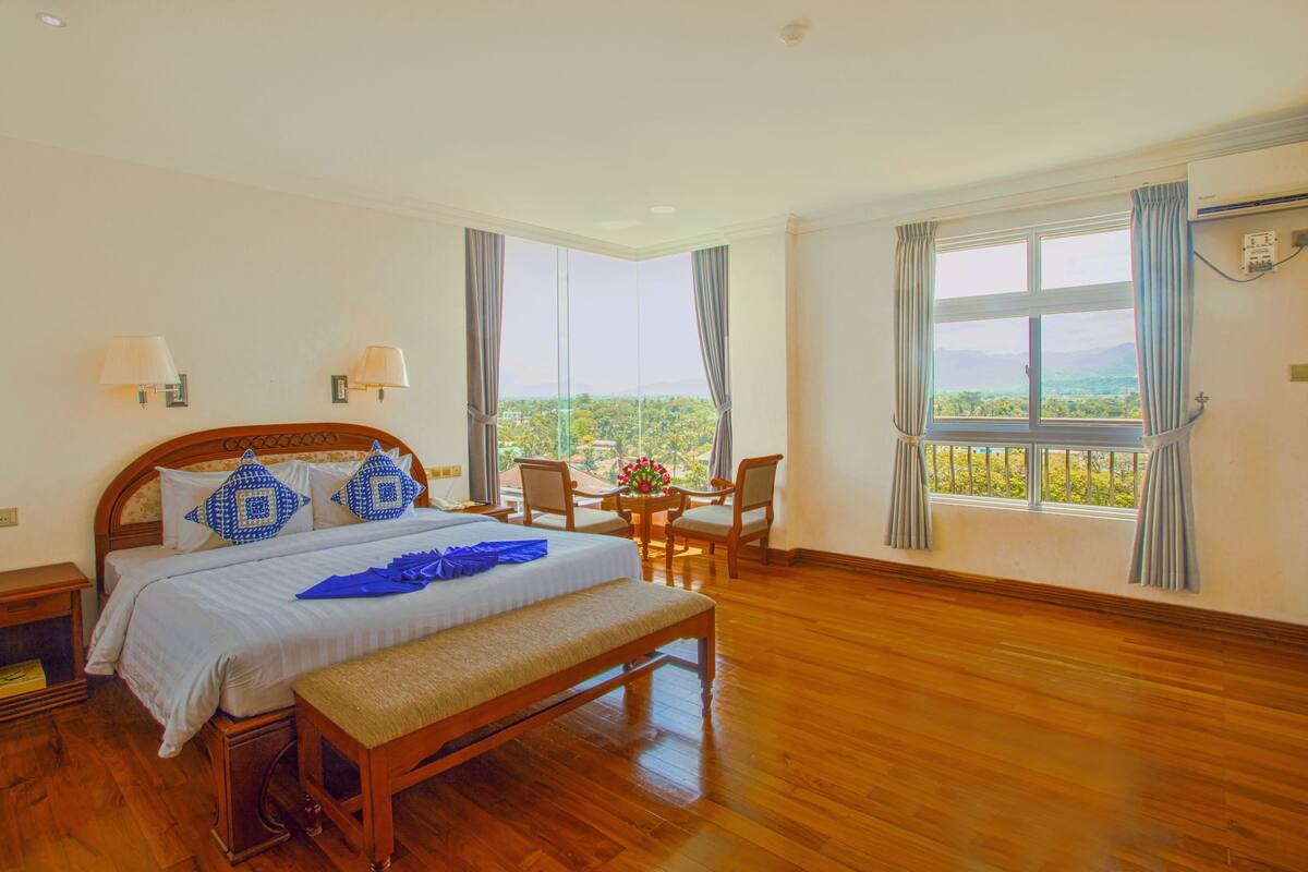 A spacious bedroom with a large bed, a wooden bench, a small table with chairs, and a view of greenery through the window.