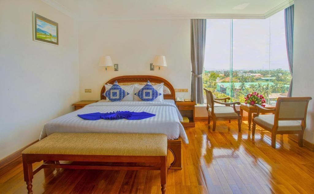A hotel room with a large bed, two bedside lamps, a wooden bench, a small table with chairs, and a view of a cityscape through large windows.