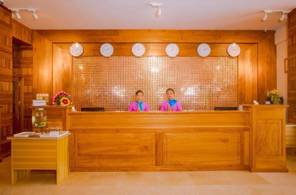 Una recepción con dos empleados, paneles de madera, relojes y una pequeña mesa con un jarrón y flores.