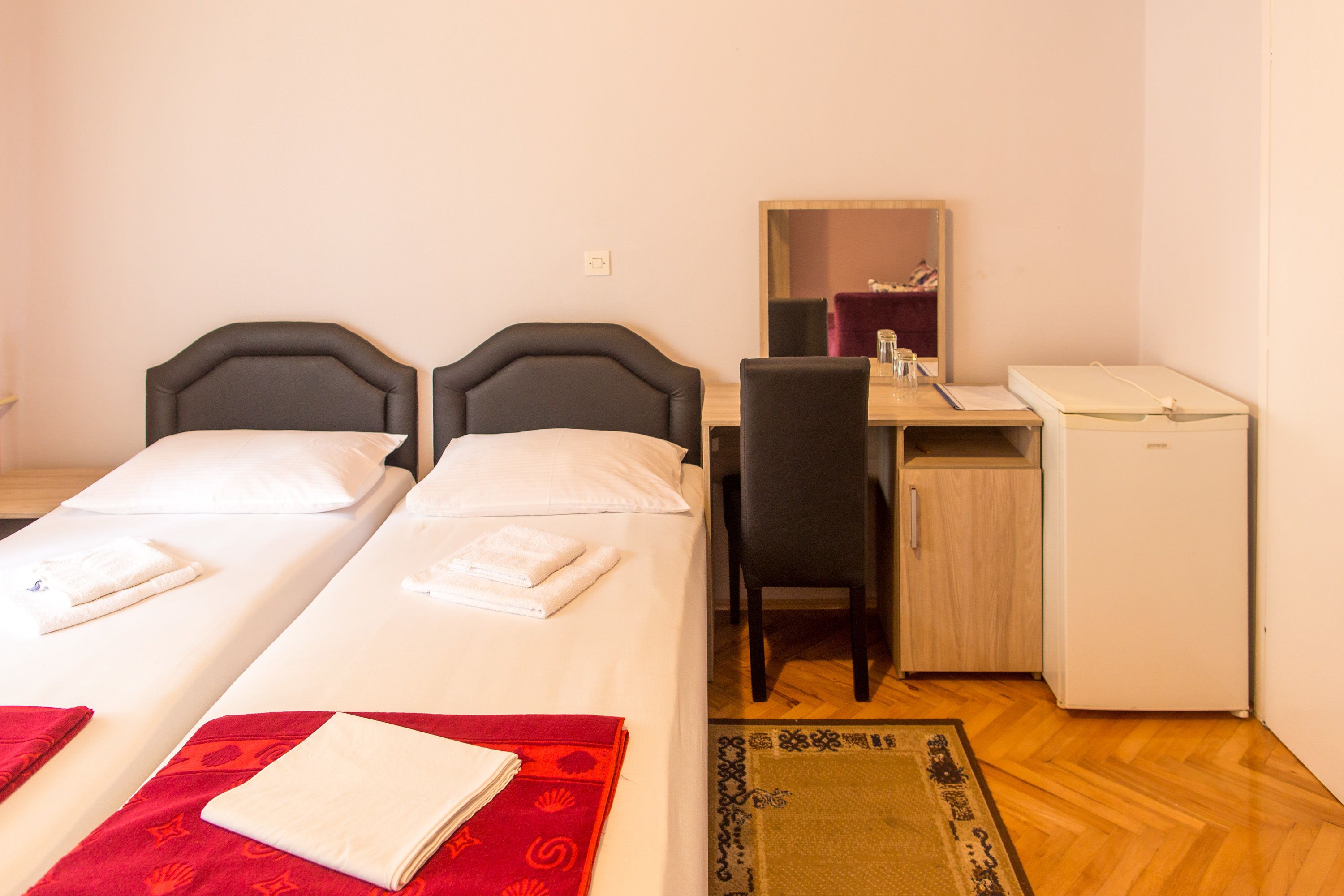A hotel room with two single beds, a desk with a chair, a small refrigerator, and a mirror.