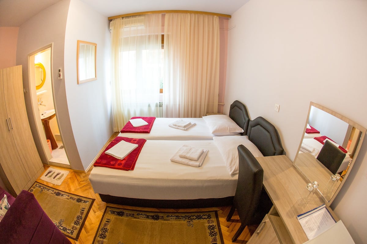 Habitación de hotel con dos camas, un escritorio, una silla, un espejo y un baño visible a través de una puerta abierta.