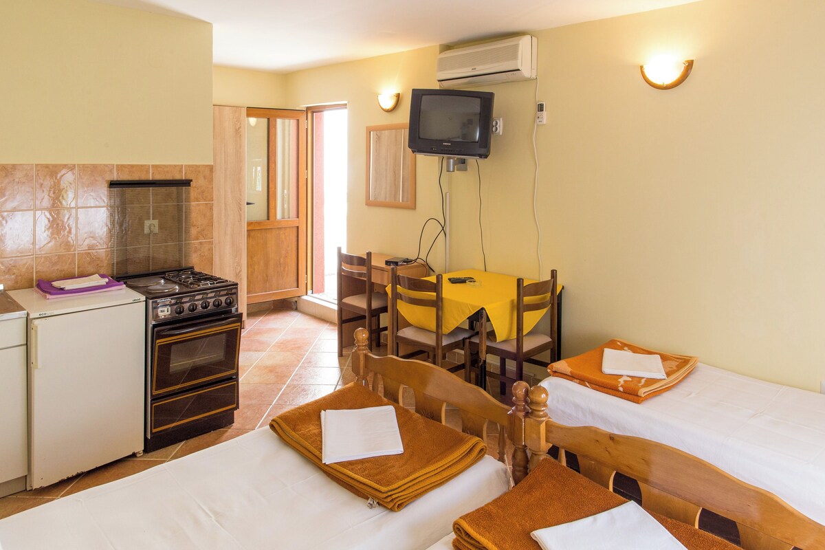 A compact kitchen with a stove, oven, and a television mounted on the wall. There are two wooden tables with chairs, a yellow tablecloth, and a door leading to another room.