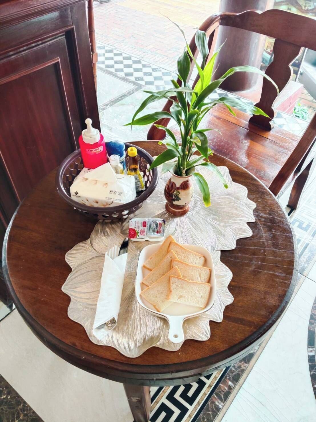 A round wooden table with a placemat, a potted plant, a bowl of bread, a bottle of ketchup, and a bottle of mustard.