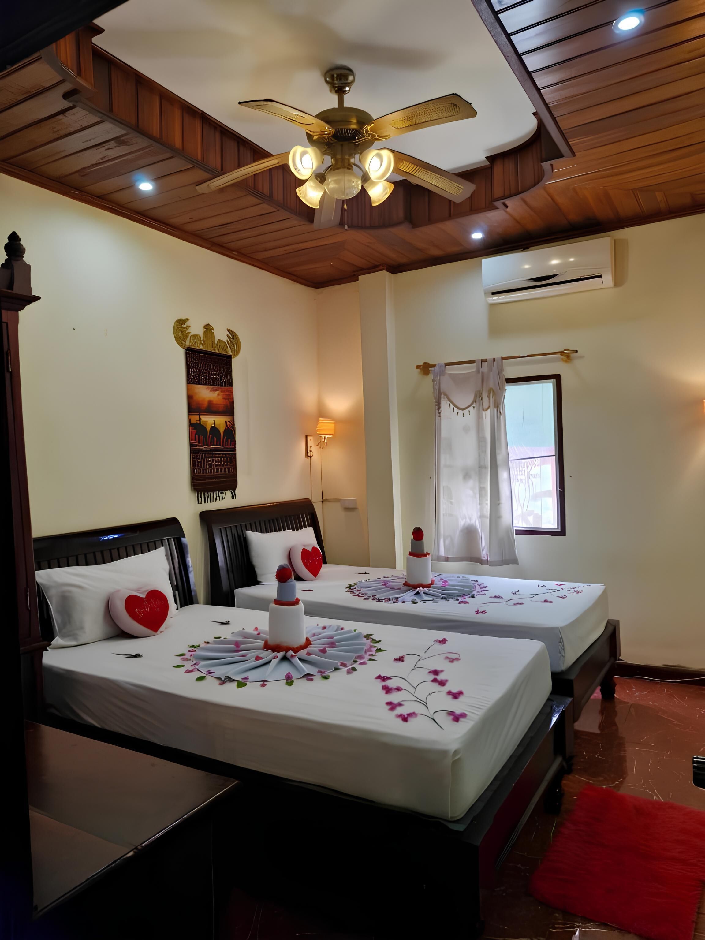 A hotel room with two beds, a ceiling fan, a wall-mounted air conditioner, and a window with curtains.