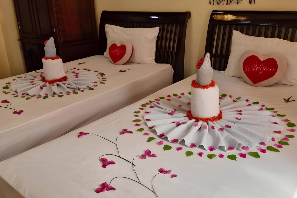 Habitación de hotel con dos camas, cada una decorada con una colcha blanca, pétalos de flores rosadas y una toalla blanca dispuesta en forma circular.