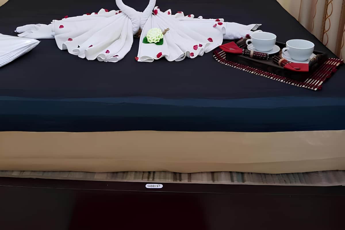 Una cama bien hecha con una colcha azul oscuro, dos almohadas decorativas rojas y una almohada decorativa marrón, y un arreglo de toallas con forma de corazón.