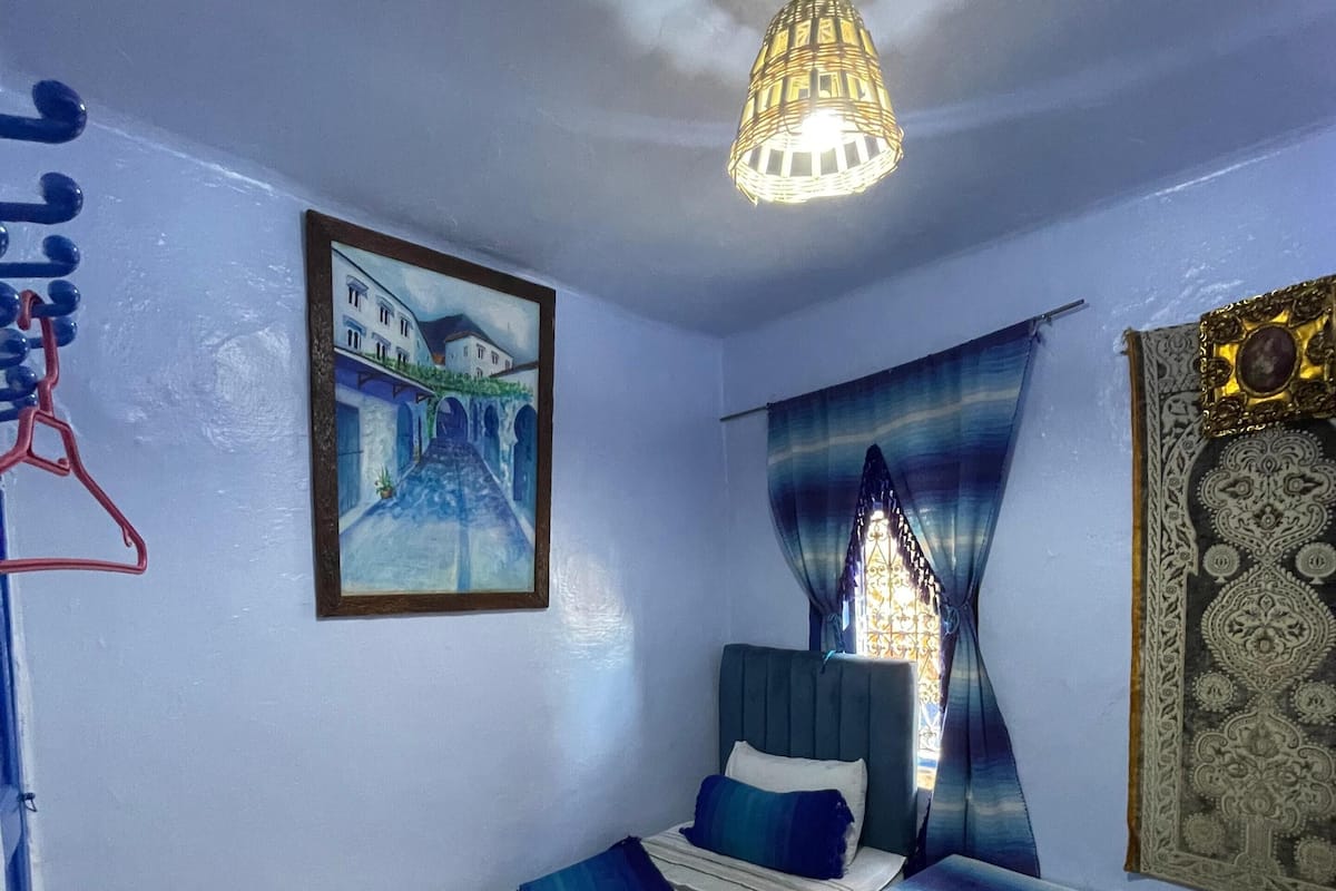 A bedroom with a bed, a bedside table, a hanging light fixture, a painting, and a patterned rug.