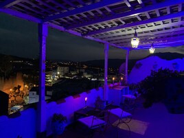 An outdoor terrace with a view of a town at night, featuring a pergola and seating area.