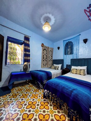 A room with two beds, a window with curtains, a patterned floor, and a hanging light fixture.