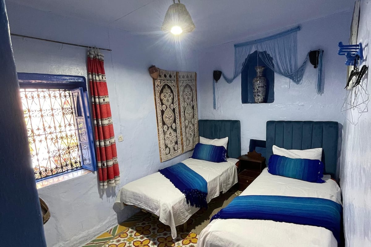 A room with two single beds, a patterned floor, a window with curtains, and a hanging light fixture.