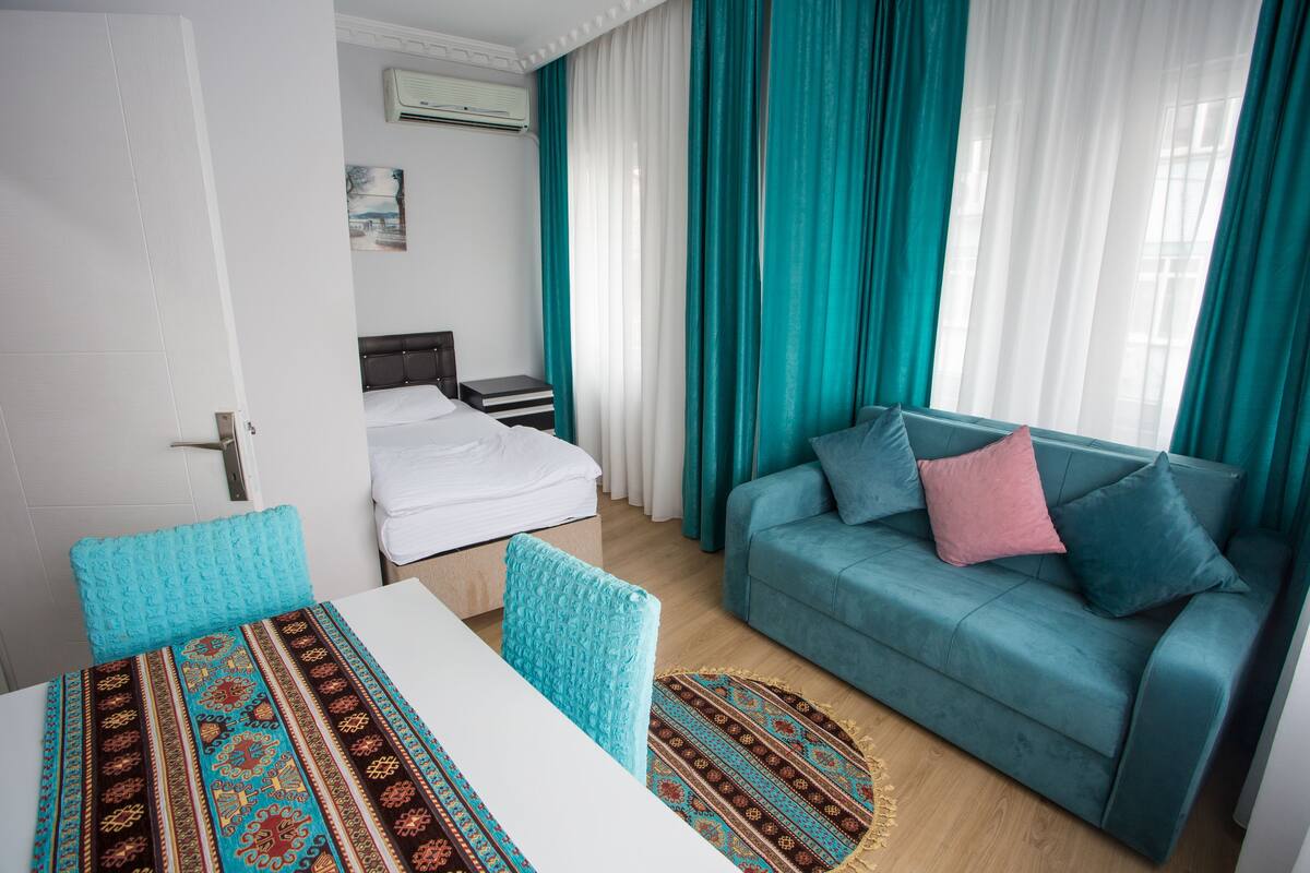 A hotel room with a bed, a blue sofa, a dining table, and teal curtains.