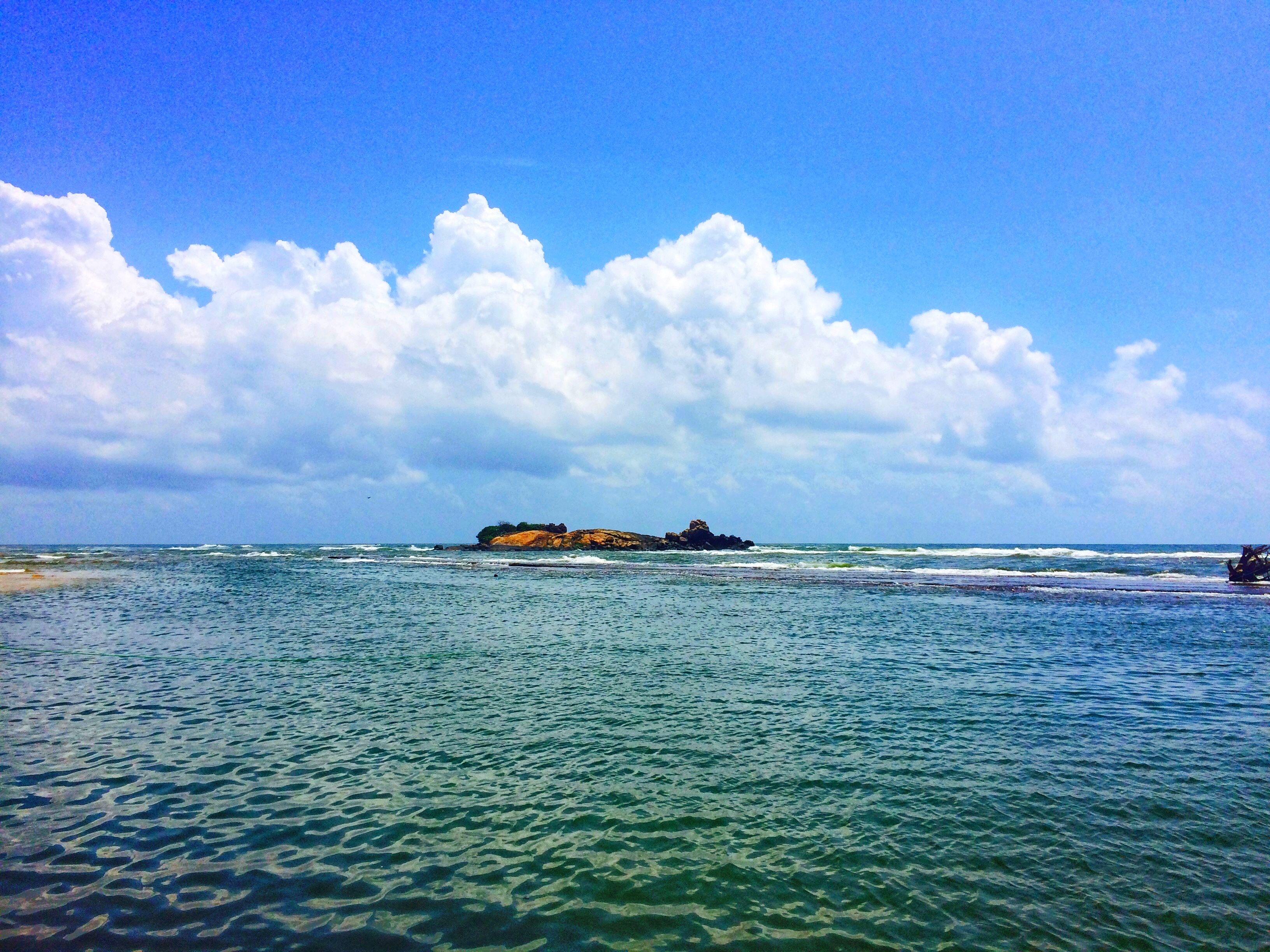 A beach with clear water, a rocky outcrop, and a partly cloudy sky.