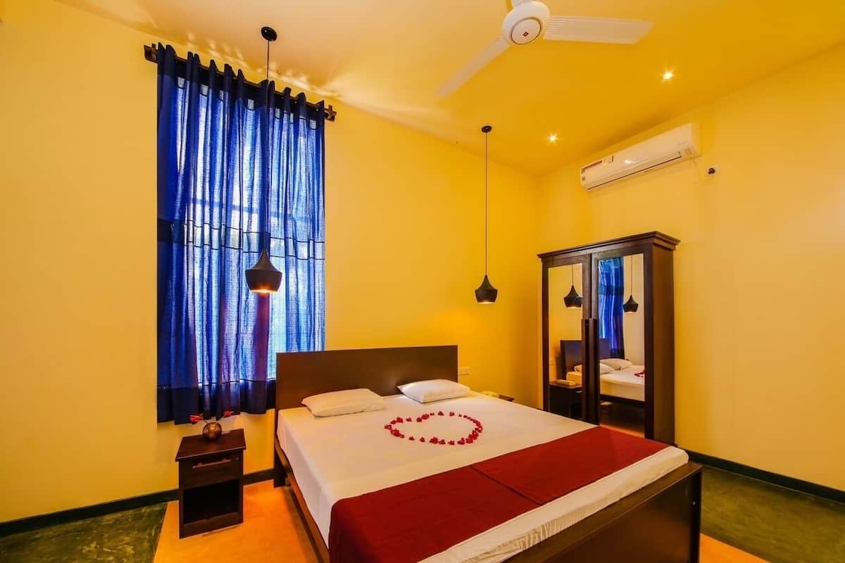 Ein Hotelzimmer mit Bett, Deckenventilator, Klimaanlage, Nachttisch, Spiegel und Fenster mit blauen Vorhängen.
